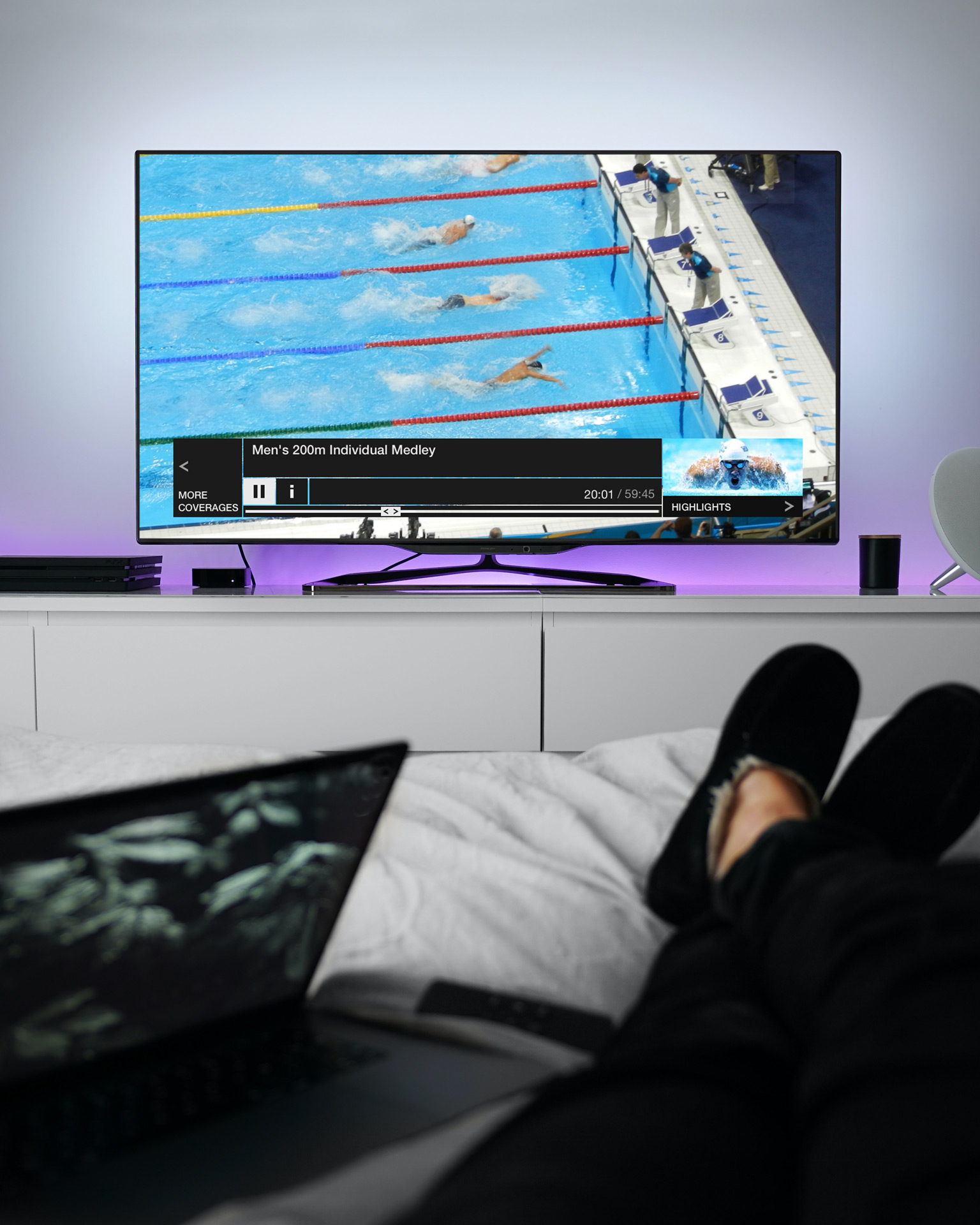 In a bedroom, there is a TV on a stand displaying a mockup of the Red Button+ live player featuring the Men's 200m Individual Medley. The coverage shows four swimmers in a pool. At the forefront of the image, a person's laptop and feet can be seen.