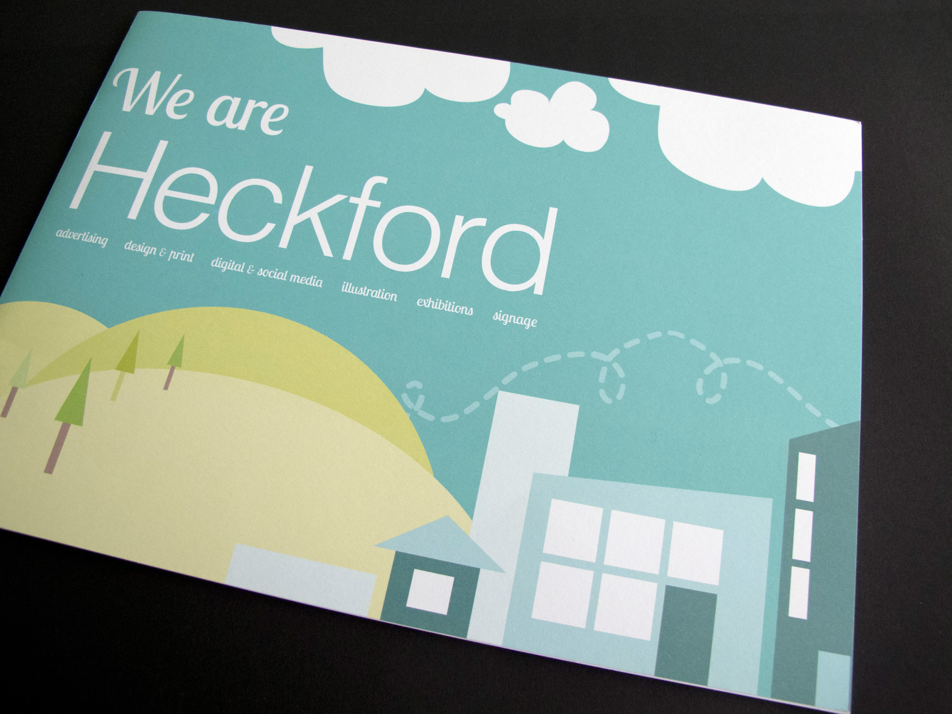 Photo of the brochure cover that depicts a vector illustration. There are green hills with trees on the left, and interlocking blue buildings on the right. There is a bright turquoise sky with white clouds and the words 'We are Heckford' in white text. The subheading lists Heckford's services.