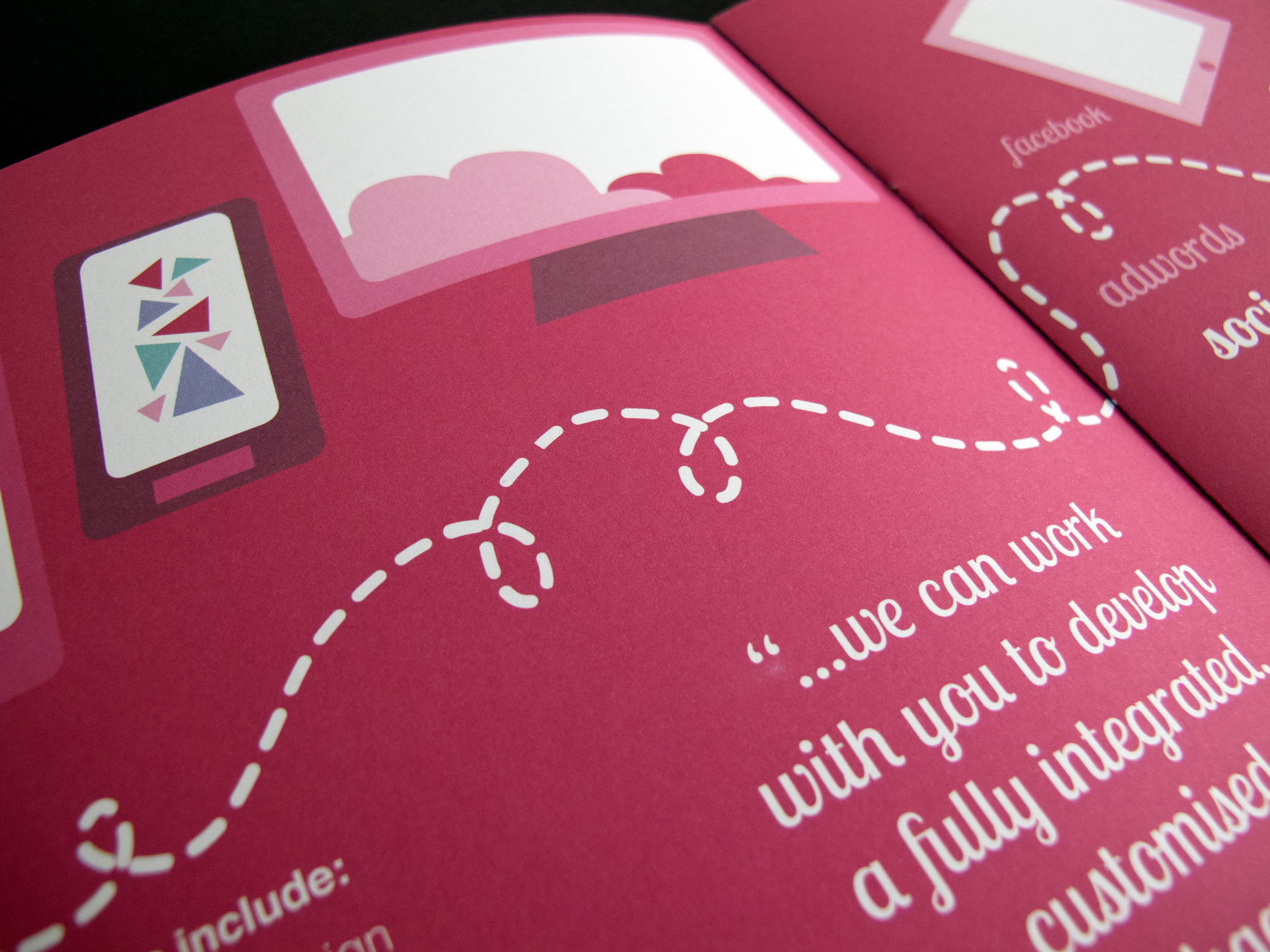 A close-up, angled photo of the centre of the digital and social media brochure page spread. The pages are a bold pink colour with white text. The vector illustration depicts mobile, desktop, and tablet devices. There is a white dotted swirly line across the page to represent connectivity.