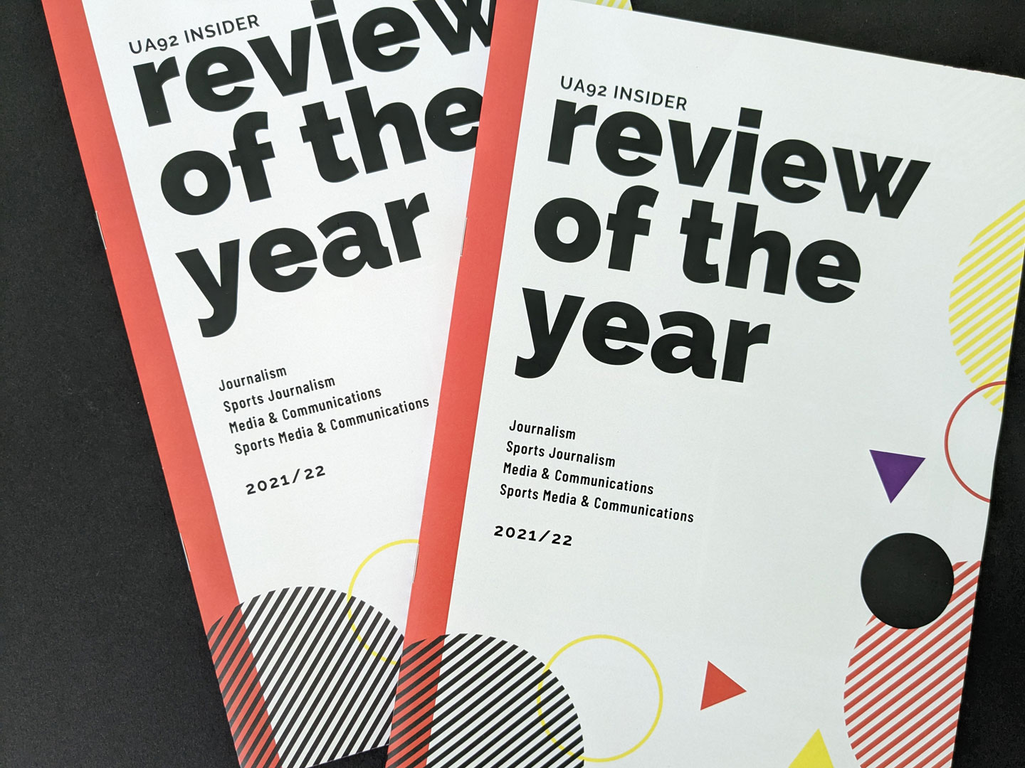 A photo of two magazines stacked on top of each other. The front cover of the magazines showcases vibrant colours, bold typography, and geometric patterns. The title 'UA92: Review of the Year' is displayed prominently in a bold sans serif font on a pale grey background. The geometric elements are coloured in hues of red, yellow, purple, and dark grey.